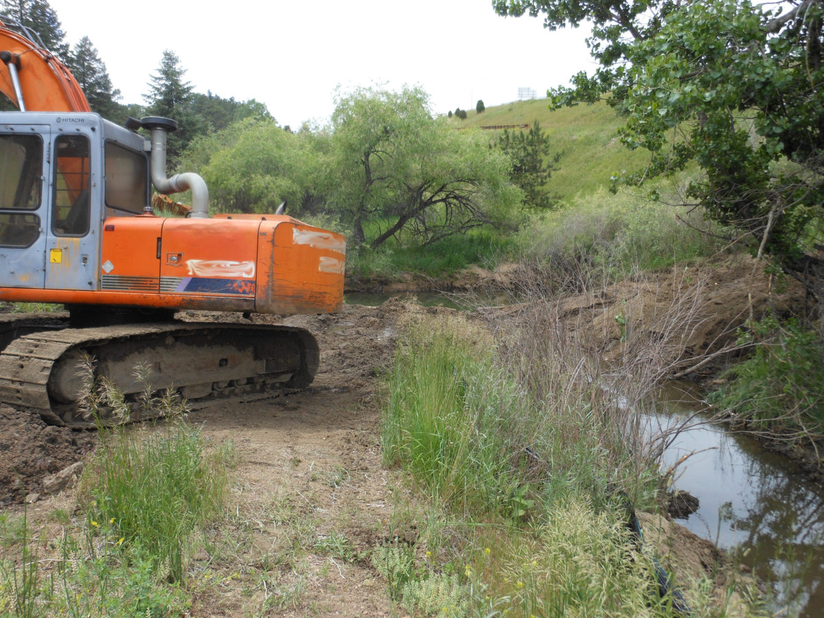 An orange excavator working near a small stream in a green, hilly area.